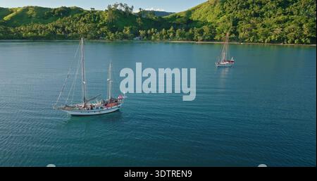 Zwei Segelboote schweben sanft auf dem tiefblauen Wasser einer tropischen Bucht, mit einer grünen, palmengesäumten Inselküste und sanften grünen Hügeln, die eine atemberaubende Kulisse unter klarem Himmel bieten Stockfoto