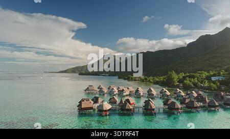 Tropische Insellandschaft mit luxuriösen Bungalows über dem Wasser, die sich in die klare türkisfarbene Lagune erstrecken, umgeben von üppigen grünen Bergen und einem wunderschönen blauen Himmel Stockfoto