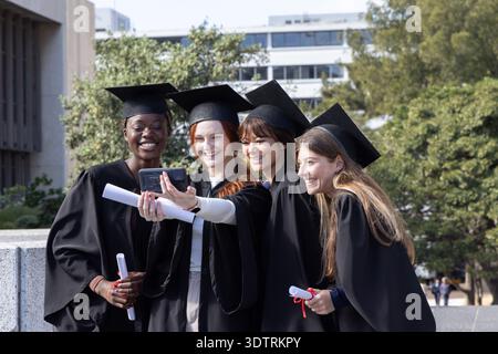 Verschiedene weibliche Absolventen machen Gruppenselfie mit Telefon auf dem Campus in Umhang und Mützen, mit Diplomen Stockfoto