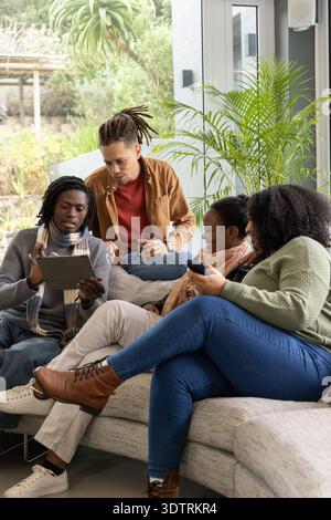 Verschiedene Freunde sitzen auf dem Sofa in der Nähe der Glasschiebetür, halten Tablet und Smartphone. Gruppe, Gerät, Freizeit, Lifestyle, Innenraum, Tageslicht, modern Stockfoto