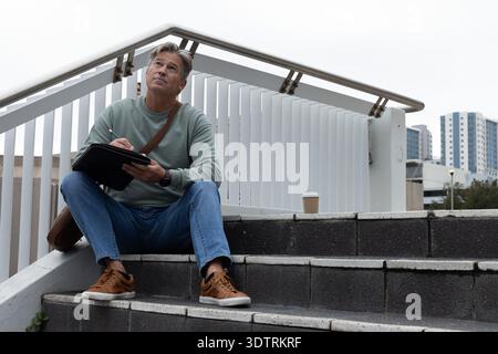 Mann mittleren Alters, der auf Betontreppen sitzt, in schwarzem Folio mit Stift schreibt, Kaffeetasse in der Nähe Stockfoto