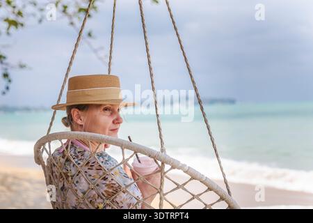Frau mittleren Alters, die sich im Hängesessel am tropischen Strand entspannt, kaltes Getränk hält, Ozeanwellen und bewölkter Himmel im Hintergrund, ruhiger Reisestil Stockfoto