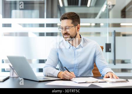 Junger männlicher Profi in Brille und blauem Hemd, der an einem modernen Schreibtisch sitzt, einen Laptop benutzt und auf Papiere schreibt, was Hingabe und hektische Arbeit widerspiegelt Stockfoto