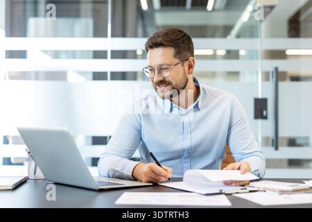 Junger bärtiger Geschäftsmann in Brille an einem hellen, modernen Schreibtisch, der sich auf Laptop und Papierkram konzentriert und Notizen schreibt, zuverlässige, produktive professionelle Planungsberichte und Datenanalysen Stockfoto