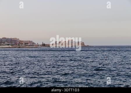 Blick auf das ruhige, tiefblaue Meer, das auf die ferne, sonnendurchflutete Stadtlandschaft trifft, schafft einen ruhigen Kontrast von Natur und Architektur, St. Julian's, Malta. Stockfoto