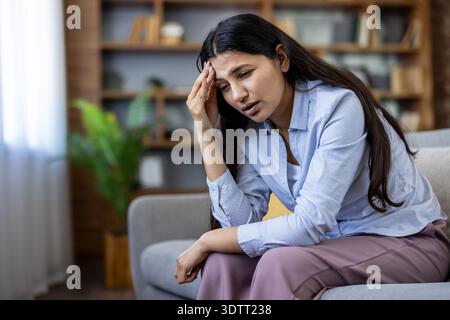 Junge Frau mit dunklem Haar, die zu Hause auf einem Sofa sitzt, ihre Stirn hält und von Migräne oder starken Kopfschmerzen zuckt, müde, gestresst und emotional ausgelaugt Stockfoto