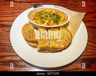 Goldener, sprudelnder gebackener Krabben-Dip mit frischen Kräutern und grünen Zwiebeln, heiß in einem weißen Gericht serviert, zusammen mit knusprigen, buttrigen Knoblauch-Toastscheiben auf A Stockfoto