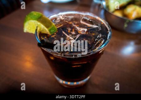 Nahaufnahme von Bourbon und Cola über Eis mit frischem Limettenkeil, serviert in einem Steinglas auf einem hölzernen Bartisch. Klassischer Whiskey-Cocktail mit sattem Bernstein Stockfoto