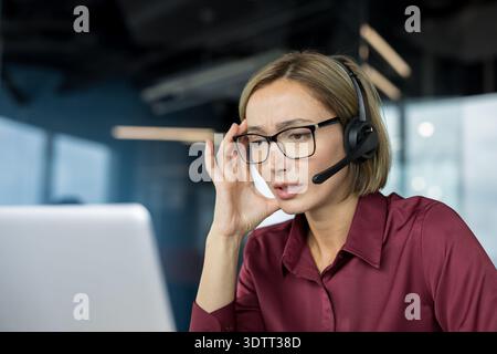 Eine gestresste Callcenter-Agentin hat schwere Kopfschmerzen und sieht müde aus, während sie in einem modernen Büro an ihrem Laptop arbeitet. Sie zeigt einen Burnout bei der Arbeit und Stockfoto