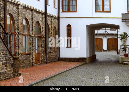 Ein alter europäischer Innenhof mit einer Mischung aus Stein- und weißen Wänden, bogenförmigen Fenstern und einem Weg, der zu großen hölzernen Scheunentüren führt Stockfoto