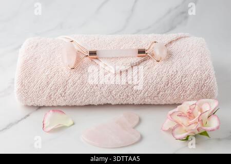 Gesichtsrolle aus Rosenquarz und Gua-Sha-Stein für Anti-Aging-Massagen mit rosa Rosenblüten und Handtuch auf dem Marmortisch. Kosmetisches Werkzeug für Stockfoto