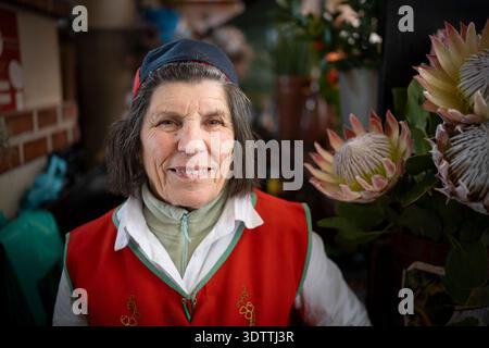 Blumenverkäuferin in traditioneller Madeira-Tracht auf dem Markt in Funchal, Madeira, Portugal, umgeben von bunten Blüten und Representin Stockfoto