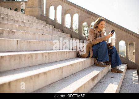 Eine Frau sitzt draußen auf Steintreppen und schaut auf ihr Smartphone. Sie hält eine Kaffeetasse in der anderen Hand. Die Sonne scheint in einem Park mit Bäumen im Stockfoto