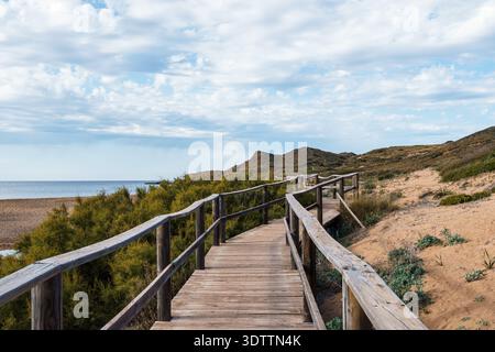 Ein malerischer Blick auf eine hölzerne Promenade, die sich durch eine sandige Landschaft zum Meer schlängelt, unter einem wunderschönen bewölkten Himmel auf Menorca. Stockfoto