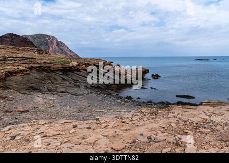 Das Bild fängt eine wunderschöne Küstenszene auf Menorca ein und zeigt mehrschichtige Felsformationen, die unter einem bewölkten Himmel auf das ruhige Meer treffen. Stockfoto