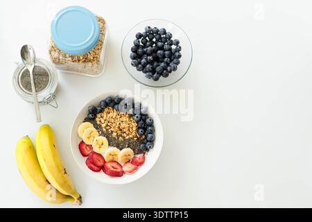 Weiße Keramikschale mit Müsli und Obst auf weißem Tisch neben Bananen, Kopierraum Stockfoto