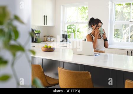 Frau, die auf der Kücheninsel steht, mit Tanktop, Becher in der Nähe von Laptop, Obstschale, Kopierraum Stockfoto