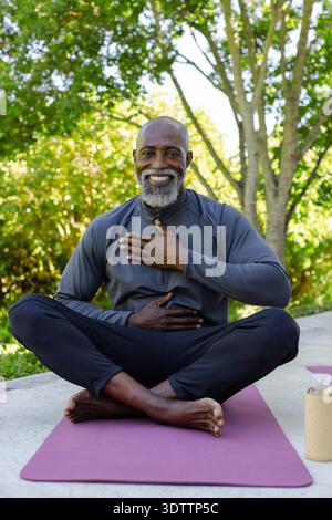 Reifer afroamerikanischer Mann atmet in sportlicher Kleidung auf lila Matte auf Terrasse mit beiger Flasche. Yoga, Pranayama, Meditation, Outdoor, Sonnenlicht, grün Stockfoto