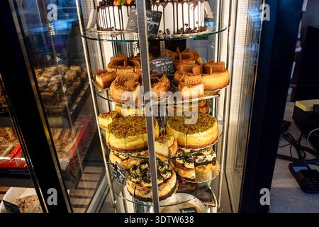 Verschiedene Käsekuchen und Kuchen in einer gekühlten Glasvitrine in einer Bäckerei oder einem Café, einschließlich Pistazien-, Oreo- und Biscoff-Sorten. Stockfoto