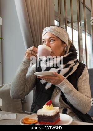 Frau trinkt Kaffee, während sie an einem Café-Tisch mit Dessertkuchen in einem modernen, gemütlichen Café-Interieur sitzt. Stockfoto