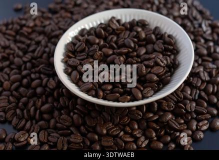 Eine saubere und professionelle Top-down-Aufnahme dunkel gerösteter Kaffeebohnen in einer weißen gerippten Keramikschale, umgeben von verstreuten Bohnen auf einer dunkelgrauen Surfa Stockfoto