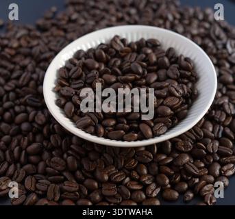 Eine saubere und professionelle Top-down-Aufnahme dunkel gerösteter Kaffeebohnen in einer weißen gerippten Keramikschale, umgeben von verstreuten Bohnen auf einer dunkelgrauen Surfa Stockfoto