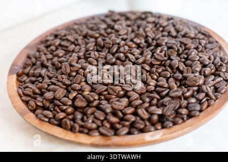Eine professionelle Nahaufnahme dunkel gerösteter Kaffeebohnen, die eine breite runde Holzschale füllen, die aus einem Seitenwinkel auf eine helle Marmoroberfläche geschossen wird Stockfoto