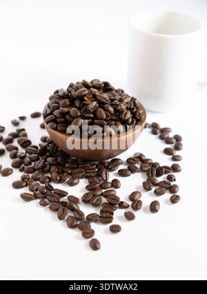 Eine hochwertige, helle Aufnahme einer kleinen Holzschale, die mit dunkel gerösteten Kaffeebohnen überflutet wird, begleitet von einem verschwommenen weißen Keramikbecher im Hintergrund Stockfoto