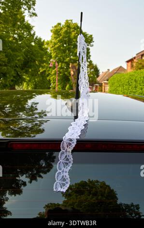 Eine vertikale Nahaufnahme des Hecks eines glänzenden schwarzen Autos, verziert mit einem langen, zarten weißen Spitzenband. Die Spitze ist über die Fahrzeugantenne gezogen Stockfoto