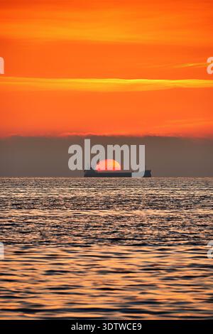 Ein beeindruckendes vertikales Foto zeigt die Silhouette eines Frachtschiffes am Horizont, das direkt vor einer großen, leuchtenden orangefarbenen Sonne als positioniert ist Stockfoto