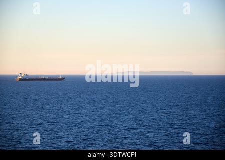 Ein Weitschuss, der ein Frachtschiff erfasst, das sich während des späten Nachmittags oder frühen Abends über das tiefblaue Meer bewegt. Der Himmelstransport Stockfoto