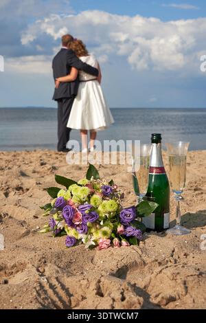 Eine romantische Aufnahme eines verschwommenen Paares, gekleidet in formeller Hochzeitskleidung, umarmt auf einem Sandstrand am Meer, mit Blick auf das Wasser unter einem Teil Stockfoto