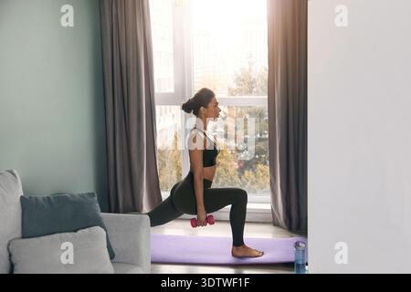 Eine Frau trainiert in Innenräumen, indem sie Ausfallschritte ausführt. Sie benutzt Kurzhanteln und steht auf einer Yogamatte vor einem Fenster. Stockfoto