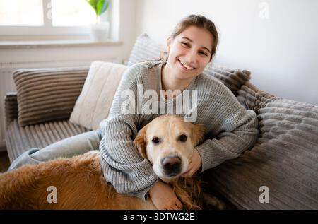 Das Mädchen sitzt auf der Couch und umarmt ihren Hund. Beide sehen glücklich aus in einem gemütlichen Raum mit natürlichem Licht. Stockfoto