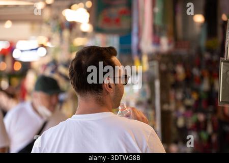 BANGKOK, THAILAND, 25. JAN 2026: Mann trinkt Wasser aus Plastikflaschen, der auf dem lokalen Markt einkaufen geht, Rückansicht, auf dem Chatuchak Markt. Stockfoto