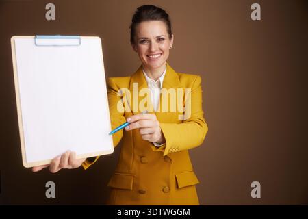Eine lächelnde Geschäftsfrau in einem gelben Blazer hält ein leeres Klemmbrett, zeigt mit einem blauen Stift darauf, führt den Zuschauer oder präsentiert Informationen Stockfoto