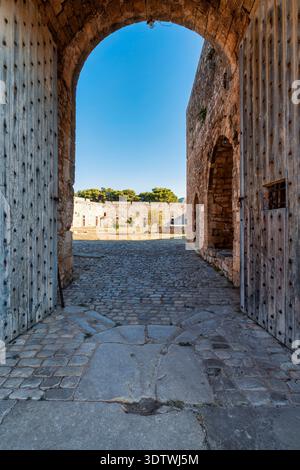 Bogengang im Inneren des Schlosses von Pylos in Griechenland, mit Holztüren, Steinmauern und einem Kopfsteinpflasterboden, der zum hellen Innenhof führt. Stockfoto