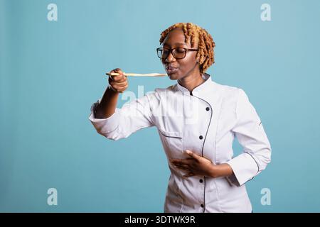 Die lächelnde Chefkochin schmeckt das Essen aus einem Holzlöffel, während sie im Studio mit blauem Hintergrund posiert. Die professionelle Köchin verkostet ihre Kreation und drückt ihre Zufriedenheit und Leidenschaft für kulinarische Handwerkskunst aus. Stockfoto