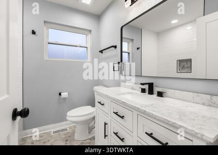 Ein modernes weißes Badezimmer mit weißem Kosmetikbereich mit Marmorablagefläche, schwarzen Armaturen, großem Spiegel und sechseckigem Fliesenboden. Stockfoto