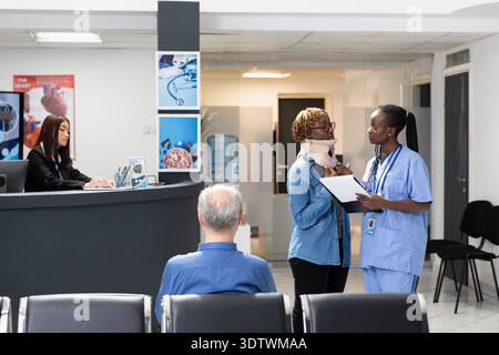 Die afroamerikanische Krankenschwester nähert sich dem Patienten mit Halsfraktur im Wartebereich der Klinik auf einen Termin. Weibliche Krankenhauspersonal hält Klemmbrett mit medizinischer Form und spricht mit Frau in der Lobby. Stockfoto