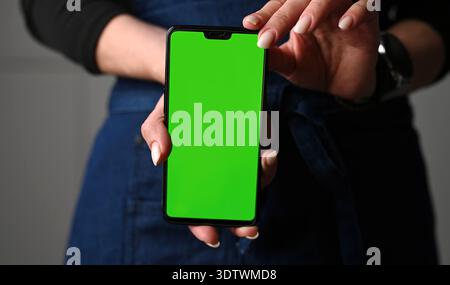 Mädchen in Schürze, das Ein Smartphone mit grünem Chromakey-Bildschirm hält Stockfoto