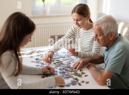 Opa und Enkelinnen arbeiten gemeinsam an Einem Puzzle, verschiedene Generationen Einer Familie verbringen Zeit miteinander Stockfoto