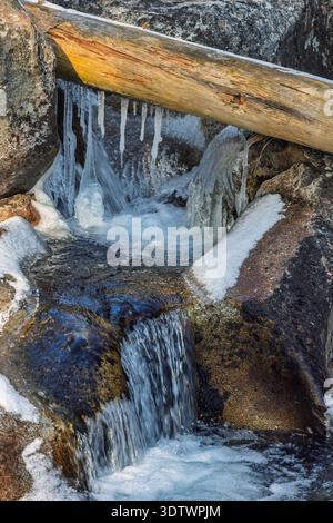 Eine fesselnde Szene eines kleinen Wasserfalls, der über Felsen stürzt, mit Schnee und Eiszapfen geschmückt, eingerahmt von einem umgefallenen Baumstamm im Winter. Hoher Ta Stockfoto
