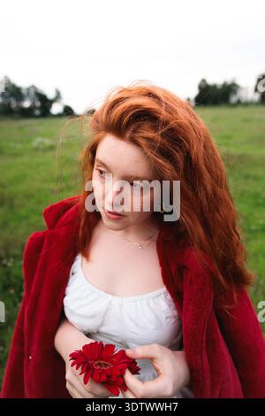 Rothaarige Frau, die rote Blume auf der Wiese hält Stockfoto