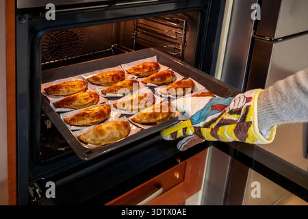 Ein Hauskoch verwendet einen Ofenhandschuh, um vorsichtig ein Tablett mit frisch gebackenen, goldbraunen Empanadas aus einem modernen Ofen zu entfernen und dabei ein köstliches hausgemachtes zu präsentieren Stockfoto