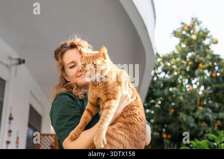 Eine Frau mit lockigen Haaren hält an einem sonnigen Tag draußen eine große Ingwerkatze in den Armen, vor dem Hintergrund eines Gebäudes und eines üppig grünen Baumes mit OP Stockfoto
