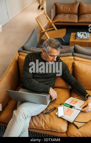 Blick auf einen älteren Geschäftsmann, der Tagebuch liest, während er im Büro auf dem Sofa sitzt Stockfoto