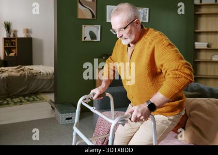 Ein älterer kaukasischer Mann, der Walker benutzt, während er im modernen Wohnzimmer steht Stockfoto