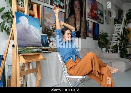 Entspannte, lächelnde Künstlerin, die Saiblinge nach einem produktiven Tag in der Loft-Studio-Wohnung streckt. Stockfoto