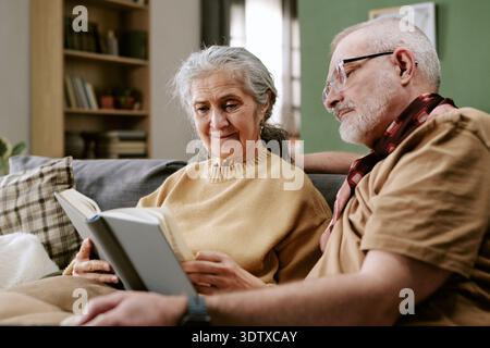 Ältere kaukasische Frau liest Buch mit einem älteren kaukasischen Mann, der sich drinnen entspannt Stockfoto